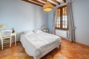 Ein Schlafzimmer mit einem Bett, einem Sessel, einem Nachttisch, einer Lampe, einem Fenster mit Vorhängen und einer Pendelleuchte.