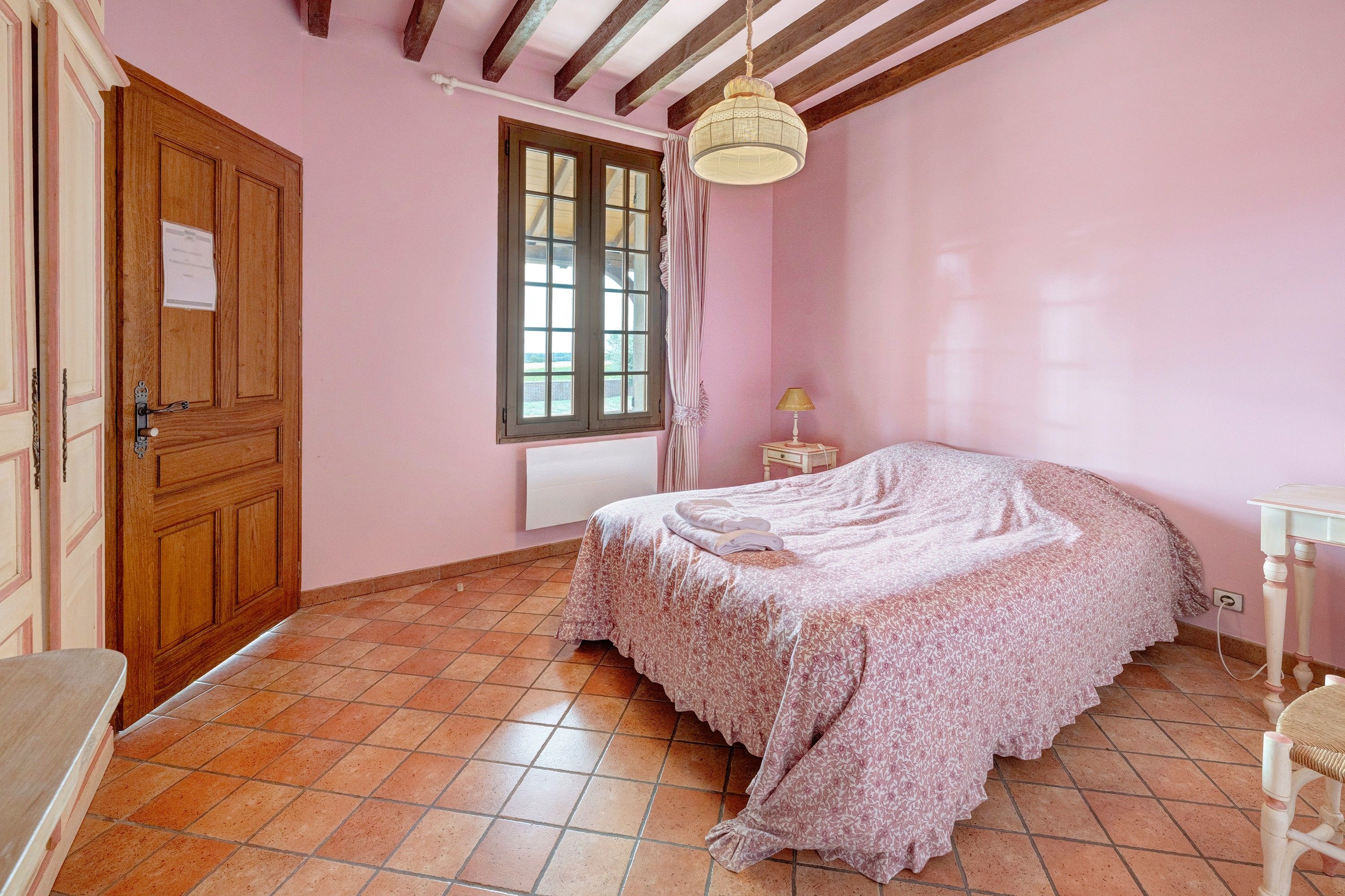 Un dormitori amb una porta de fusta, una finestra amb cortines i un llit amb llençols florals, a més d'una taula de nit amb una làmpada.