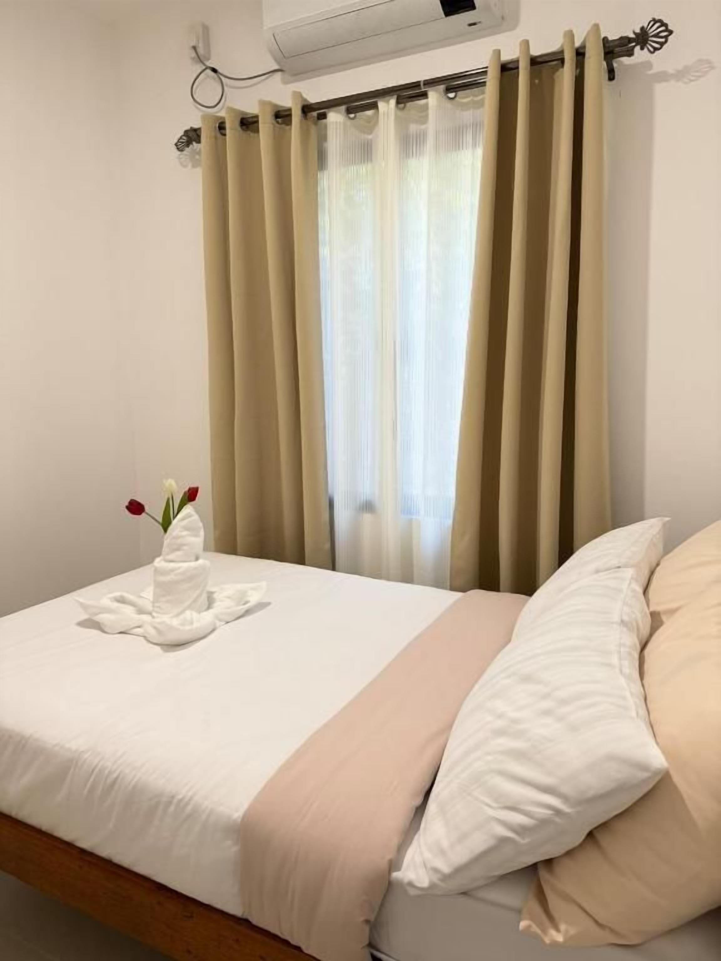 A hotel room with a bed, beige curtains, a white air conditioning unit, and a towel with flowers on the bed.