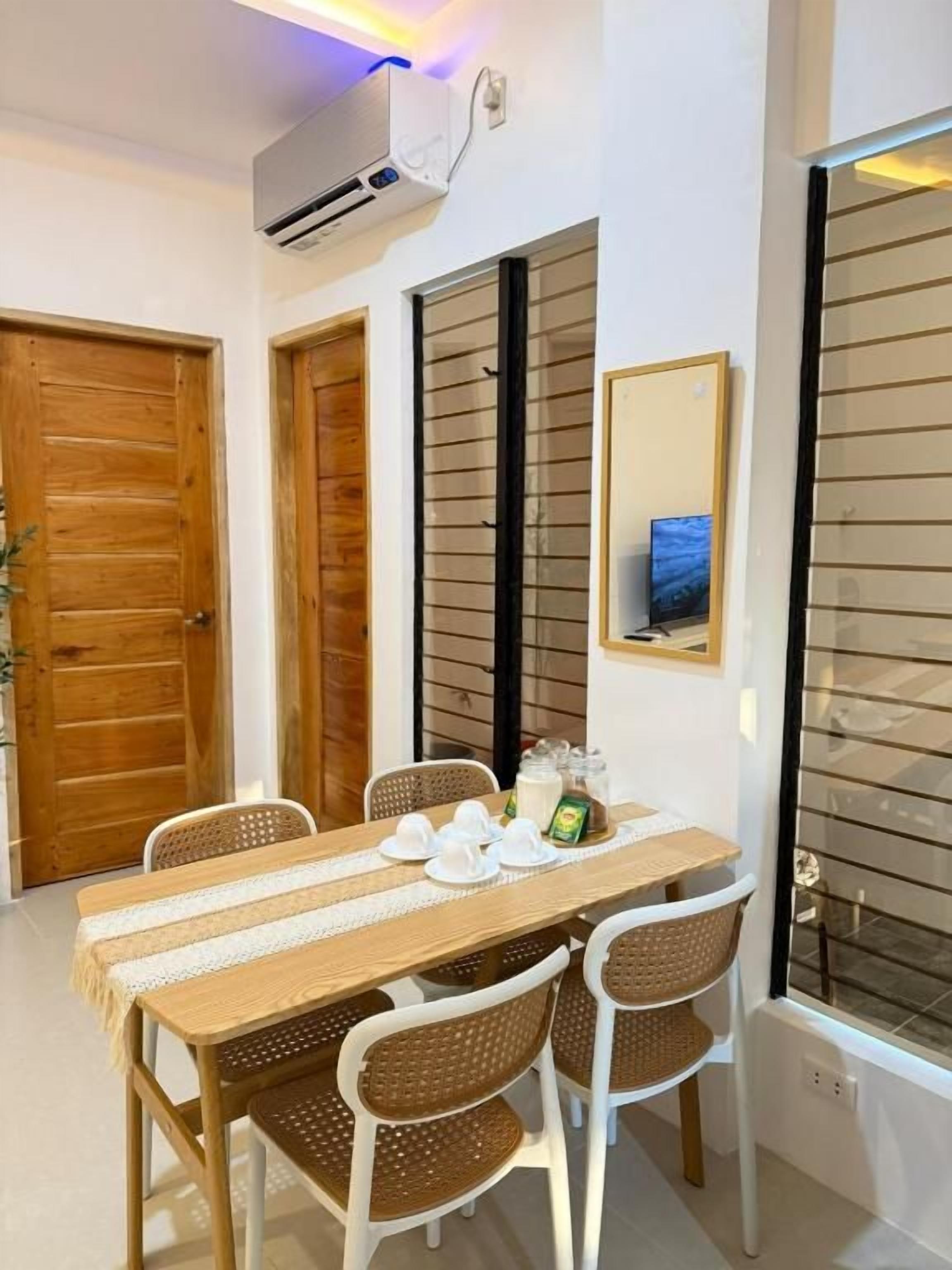 Un comedor con una mesa lista para cuatro personas, rodeada por sillas de mimbre, una mesa de madera y un equipo de aire acondicionado blanco montado en la pared.
