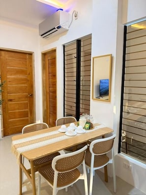 Un comedor con una mesa lista para cuatro personas, rodeada por sillas de mimbre, una mesa de madera y un equipo de aire acondicionado blanco montado en la pared.