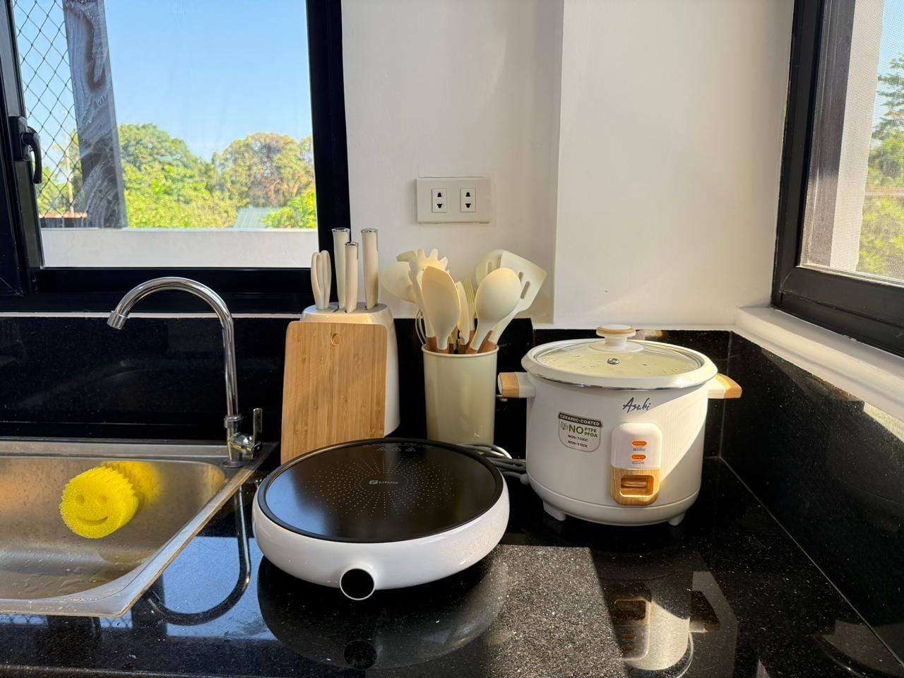 Una cocina con un lavamanos, una cocedora de arroz y un juego de palas para arroz sobre una encimera negra.