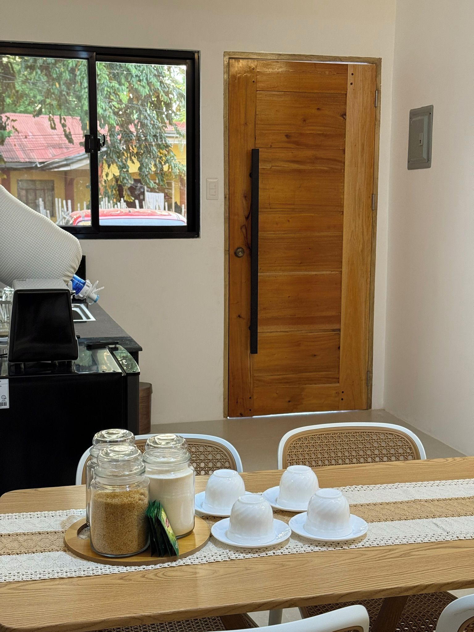 A dining table with white plates and glasses, a wooden door, and a window with a view of a red car and greenery.