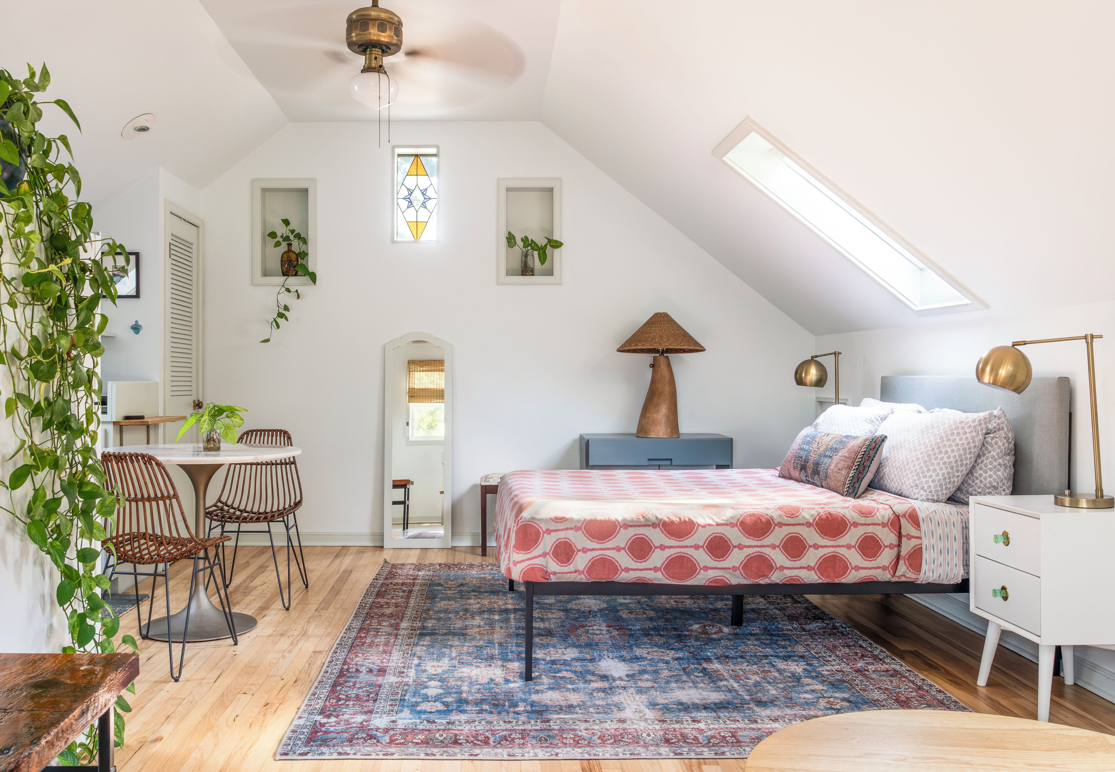 A bedroom with a sloped ceiling, a bed with patterned bedding, a desk with chairs, and a large window.