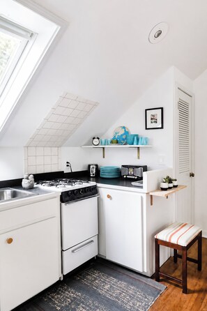 A compact kitchen with a gas stove, a toaster, and a small refrigerator. There is a window providing natural light and a door with a louvered design.