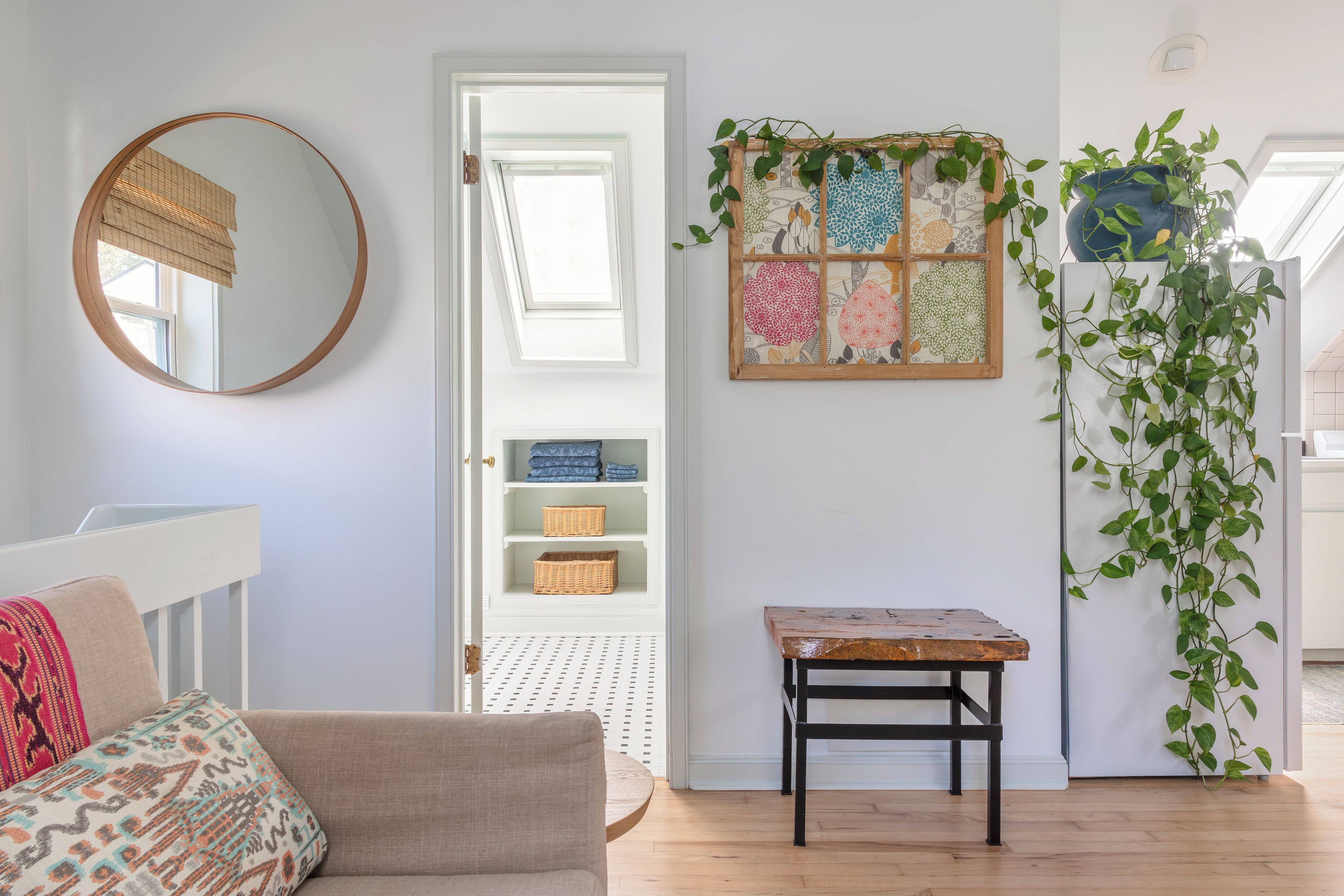 A room with a round mirror, a door leading to a storage area with wicker baskets, a small wooden table, and a wall-mounted plant.