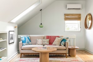 Una sala de estar moderna con un sofá, una mesa de centro redonda de madera, una mesita auxiliar, una lámpara colgante, un aire acondicionado instalado en la pared, una ventana con persianas y un espejo.