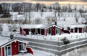 A series of red cabins with large windows and a covered patio area, set in a snowy landscape with trees in the background. - Holiday Home With 1 Bedroom (Braunlage)
