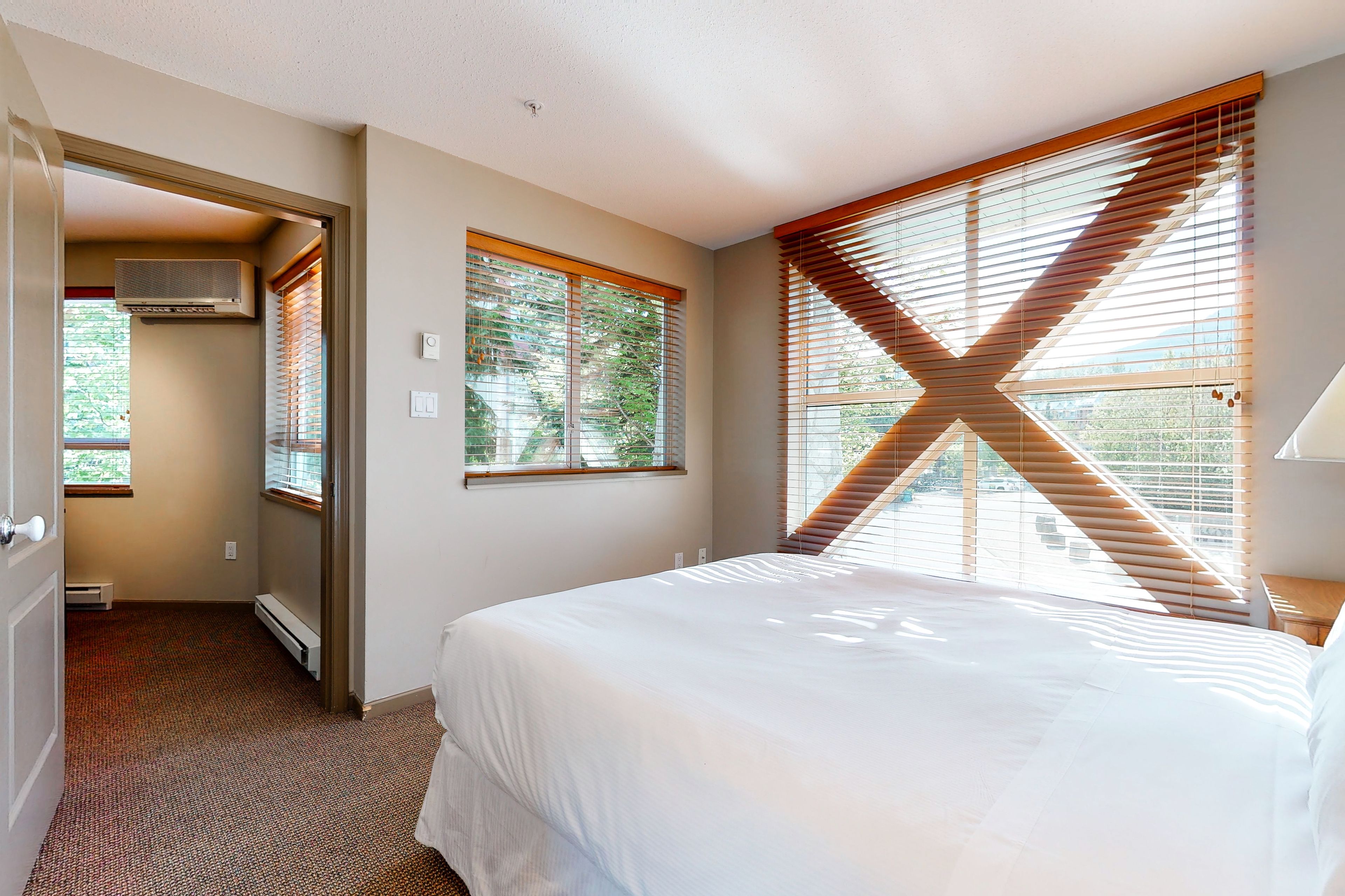 A bedroom with a large bed, two windows with blinds, a radiator, and a door leading to another room.