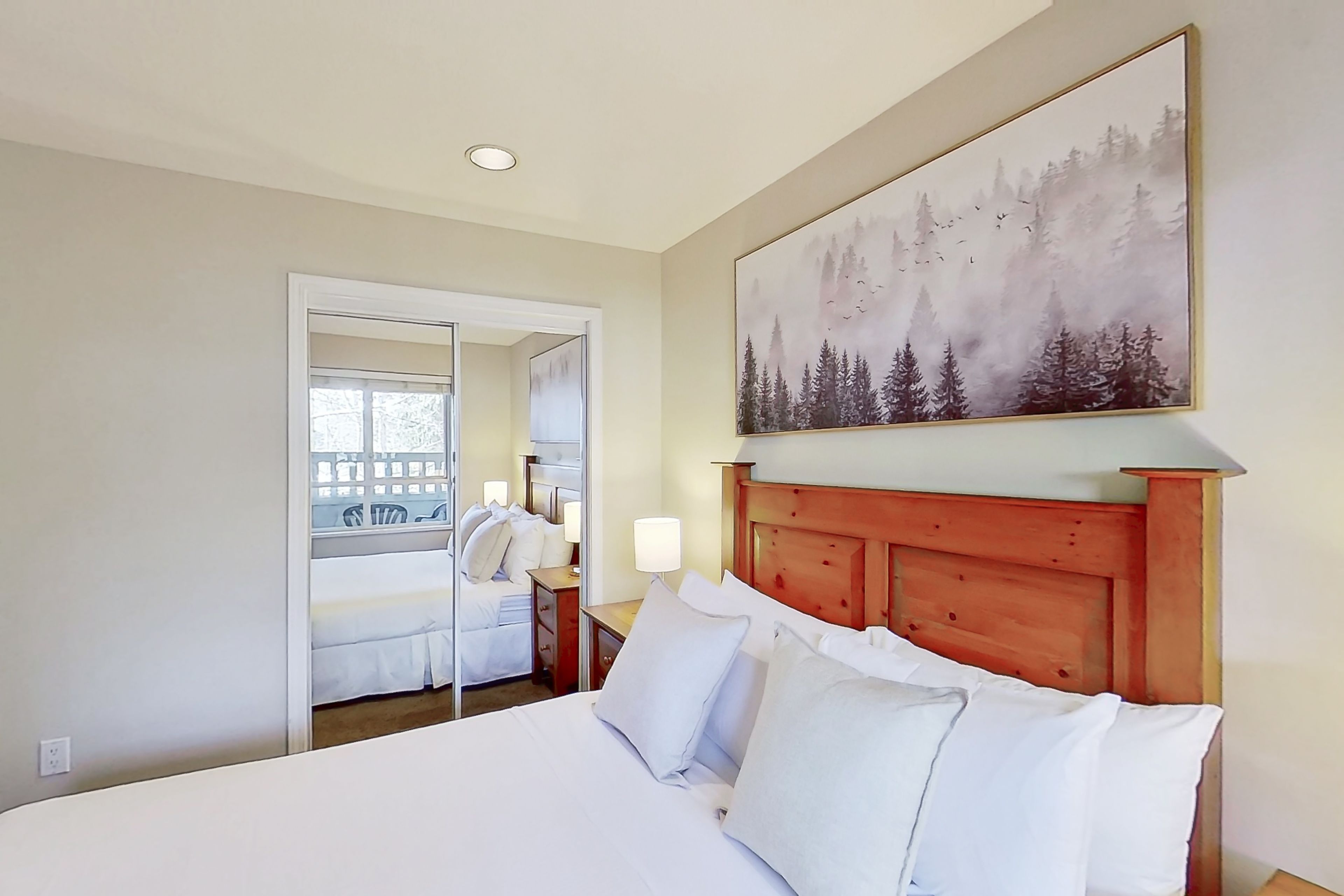 Habitación de hotel con una cama grande, cabecera de madera, un cuadro de un bosque y un espejo que refleja la habitación.