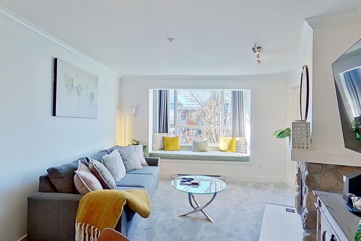 A living room with a sofa, a coffee table, a television, and a window with a view of buildings.
