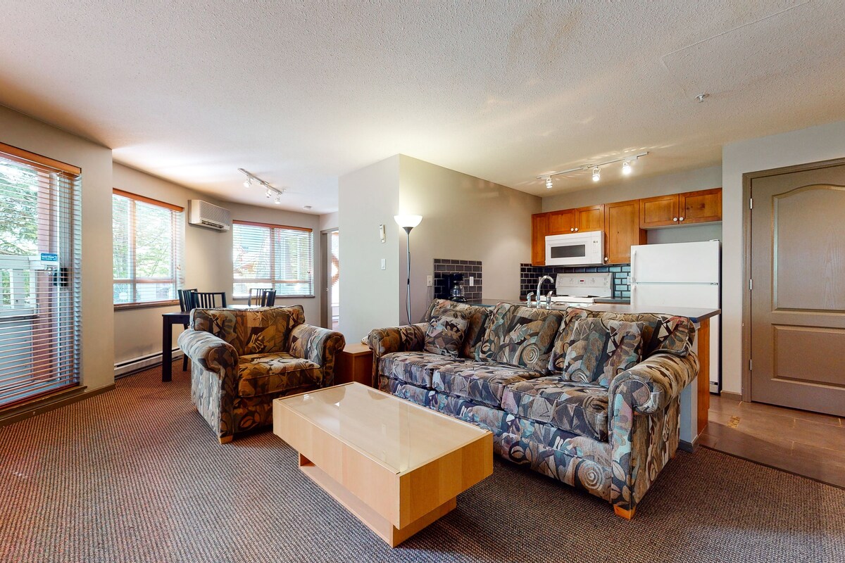 A living room with a sofa, two armchairs, and a coffee table. There is a kitchen area with a microwave and refrigerator.