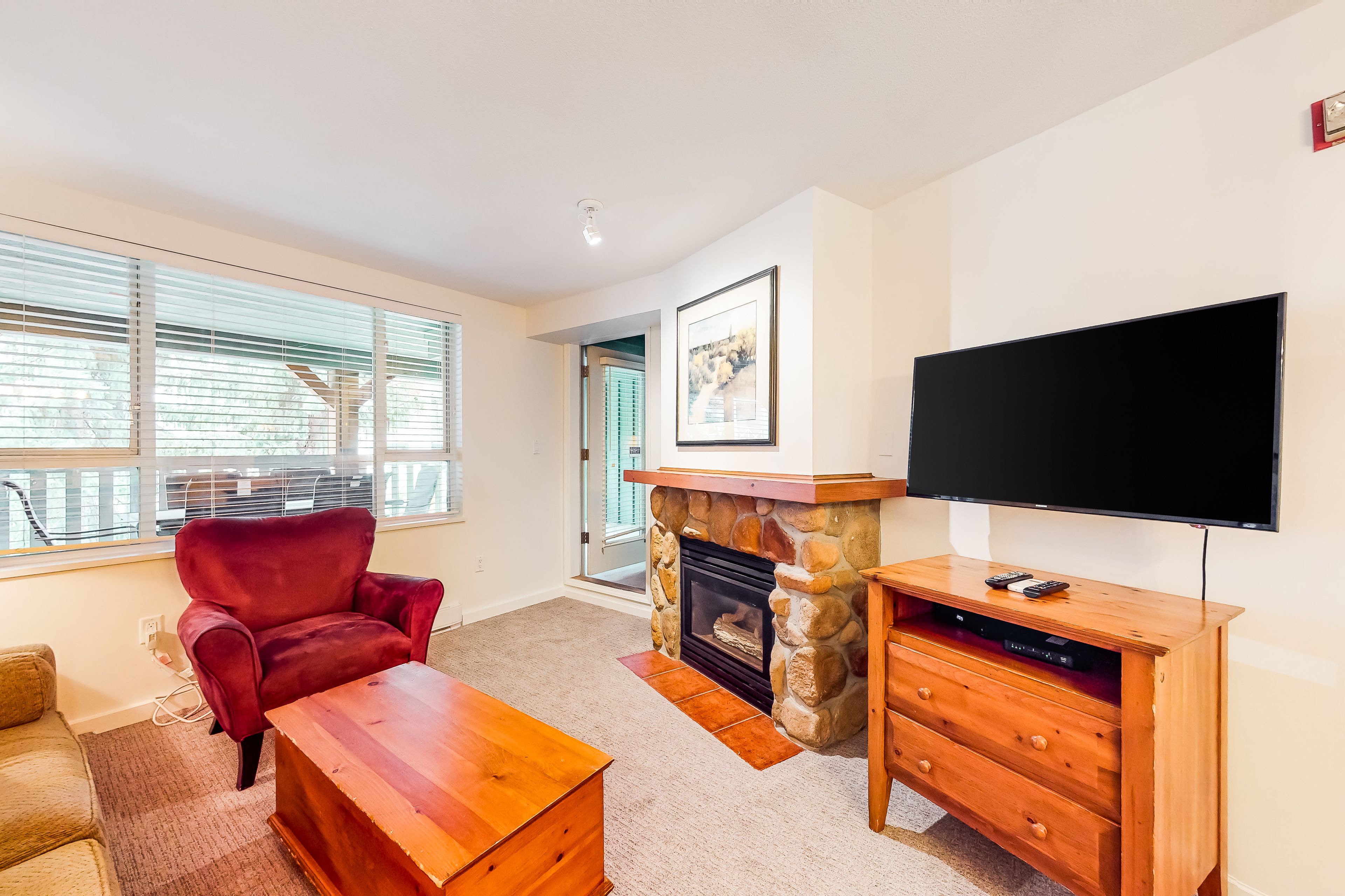 A living room with a stone fireplace, a red armchair, a wooden coffee table, and a flat-screen TV mounted on the wall.