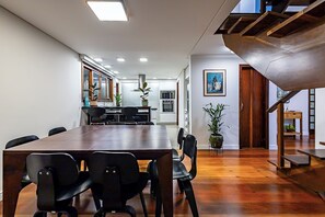 A modern dining area with a long table and chairs, a kitchen in the background, and a staircase leading to an upper level.