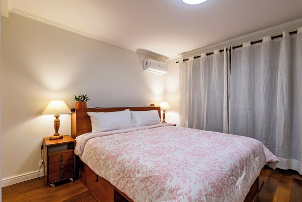 A neatly made bed with a floral bedspread, two bedside lamps, a nightstand with a drawer, and a wall-mounted air conditioning unit.