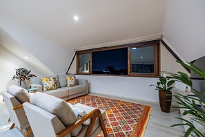 Un salon moderne avec un canapé, un tapis à motifs et une vue sur la ville la nuit, visible à travers une grande fenêtre.
