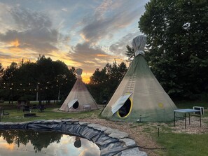 Two teepees with ladders, a small pond, and string lights in a park setting.