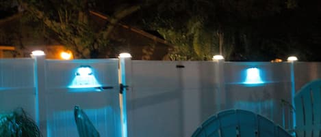 Un espace extérieur aménagé avec des chaises et un parasol, entouré d’une clôture blanche agrémentée de guirlandes lumineuses.