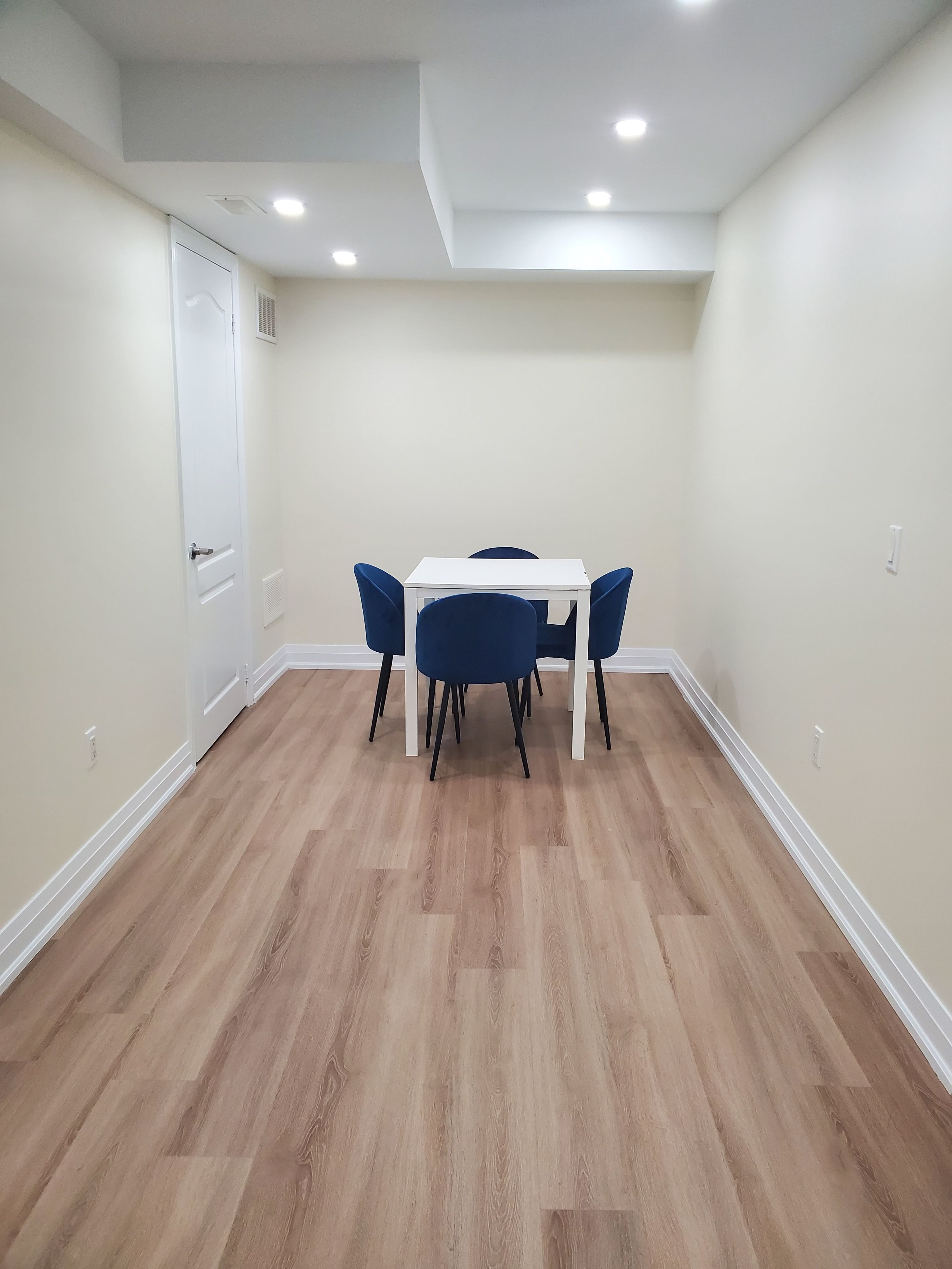 A room with a white table and four blue chairs, wooden flooring, and recessed ceiling lights.
