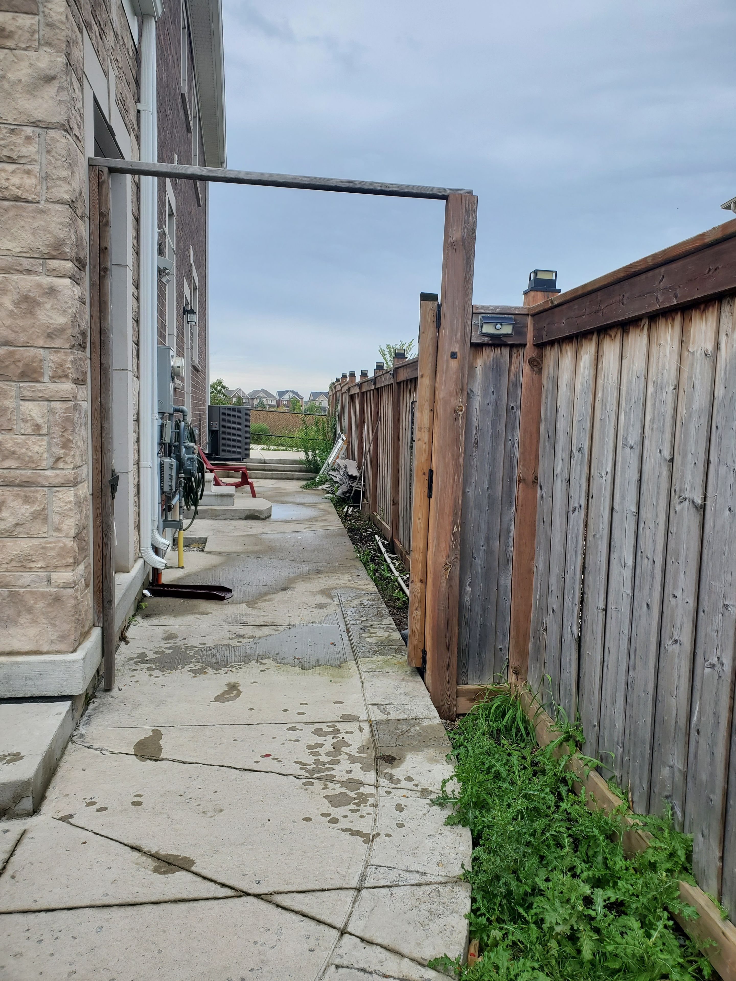 A concrete pathway leading to a wooden fence, with a building on one side and a clear sky above.