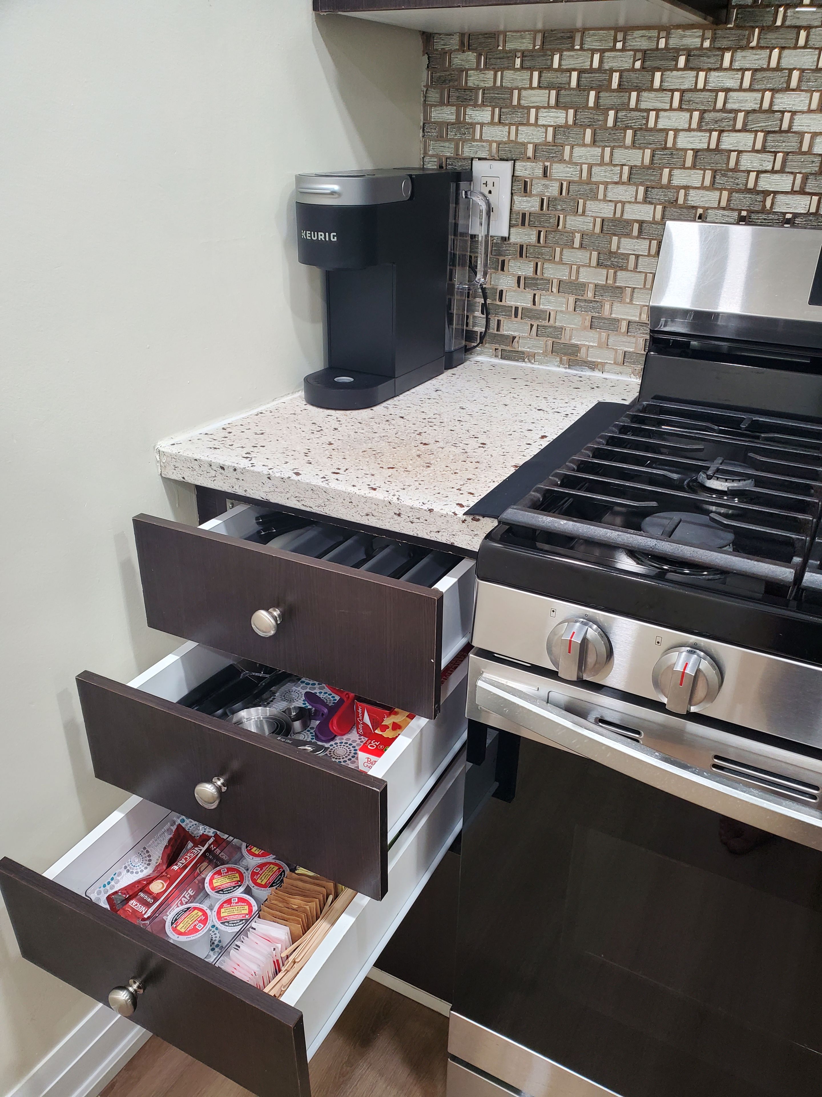 A kitchen with a coffee maker, a stove, and drawers containing kitchen utensils and packaged food.