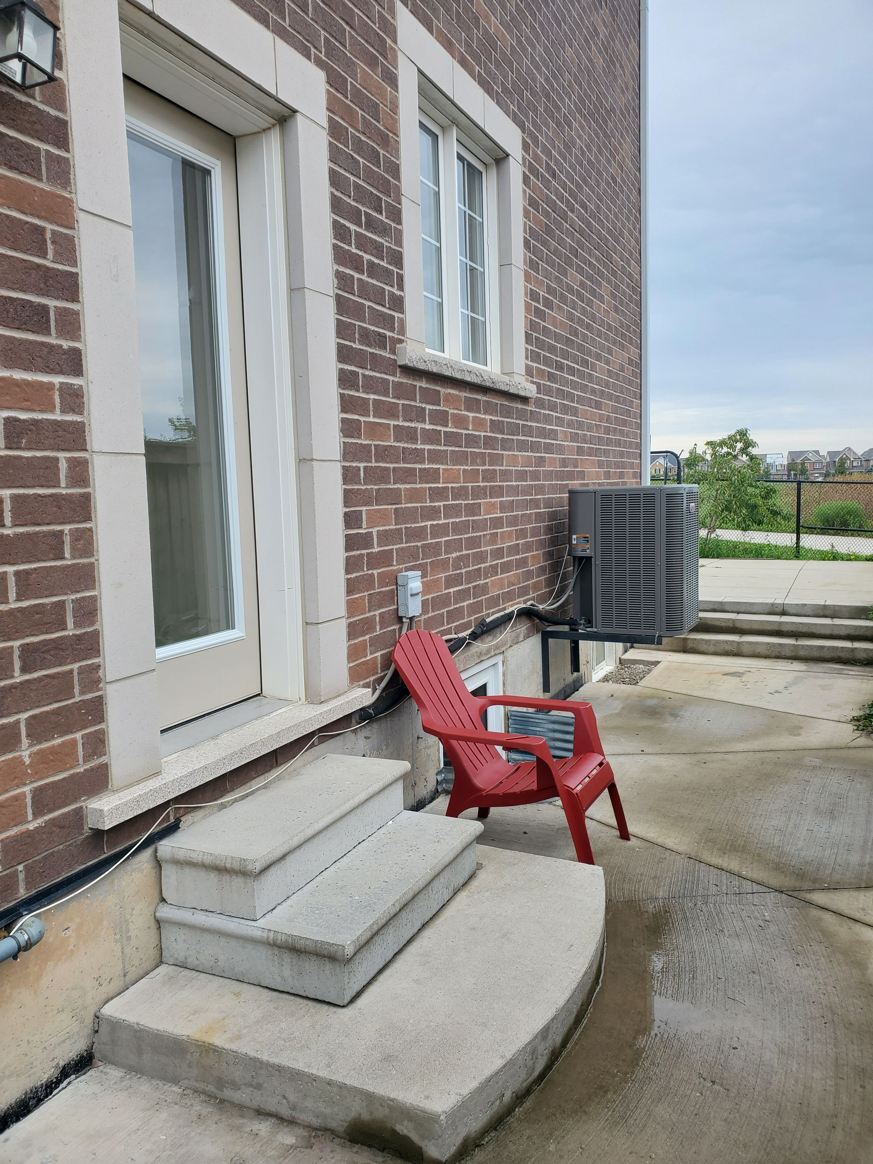Un bâtiment en briques avec une chaise rouge à l’extérieur, un escalier en béton et un appareil de climatisation.