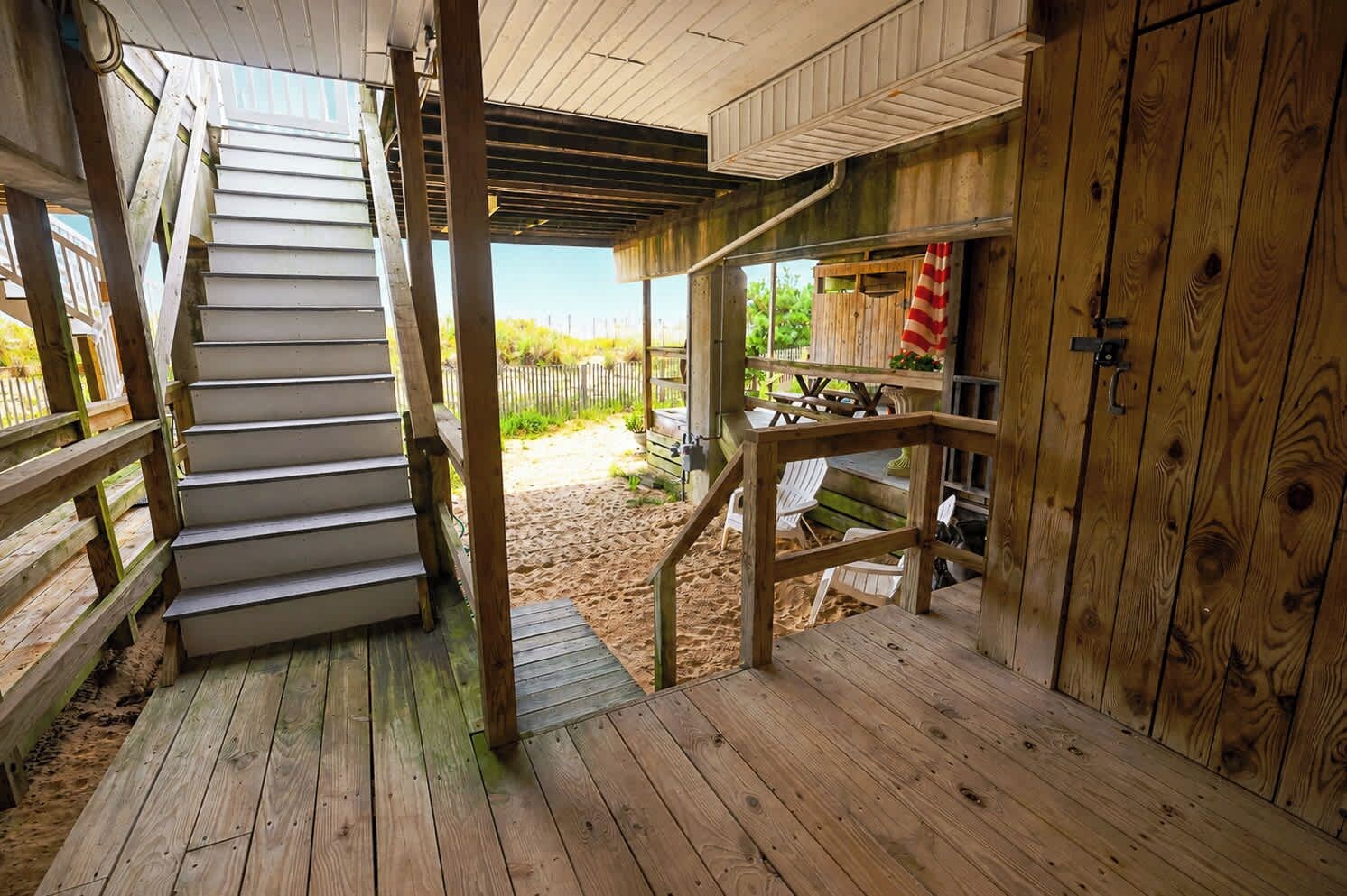 Eine Holztreppe führt zu einem überdachten Außenbereich mit Sandboden, einer Holzdielenterrasse und einem Blick auf einen Zaun im Hintergrund.