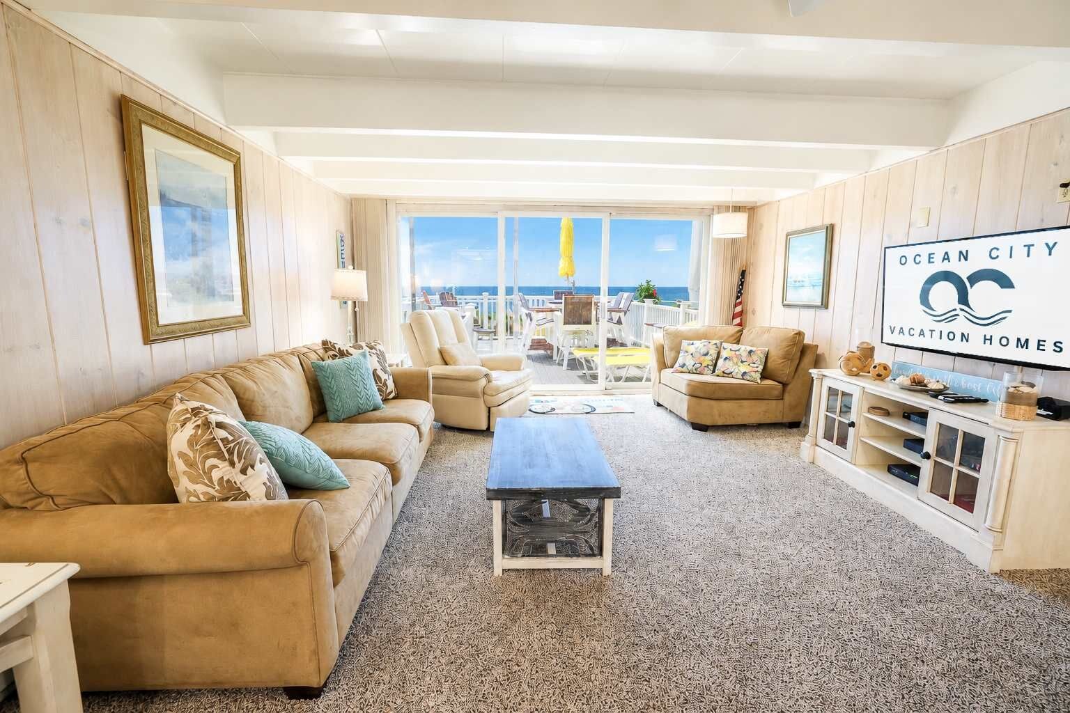 A spacious living room with a large window offering a view of the ocean, a beige sofa, a coffee table, and a TV on a wooden cabinet.