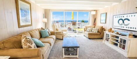 A spacious living room with a large window offering a view of the ocean, a beige sofa, a coffee table, and a TV on a wooden cabinet.