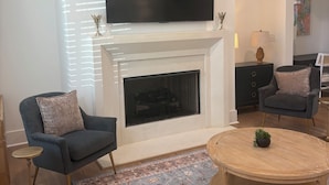 Ein Wohnzimmer mit Kamin, einem flachbildfernseher darüber, einem runden Couchtisch und einem gemusterten Teppich.