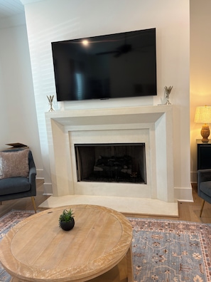 Una sala de estar con un televisor de pantalla plana montado sobre una chimenea blanca, un sofá oscuro con un cojín, una mesa de centro redonda con una planta y una alfombra estampada.