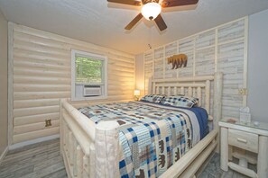 Een slaapkamer met een houten wand, een bed met een blauw-witte geruite spreidek en een plafondventilator, plus een raam met zonwering.
