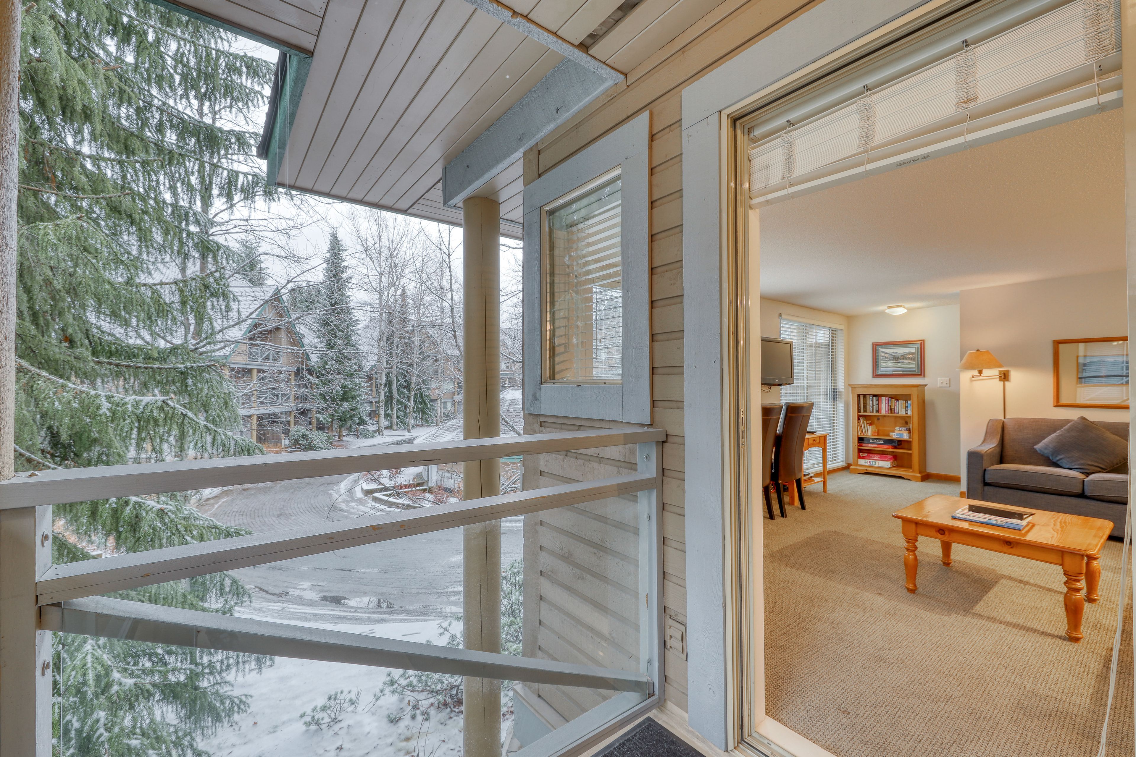 A cozy living room with a sofa, coffee table, and bookshelf. A large window offers a view of snow-covered trees and a house.