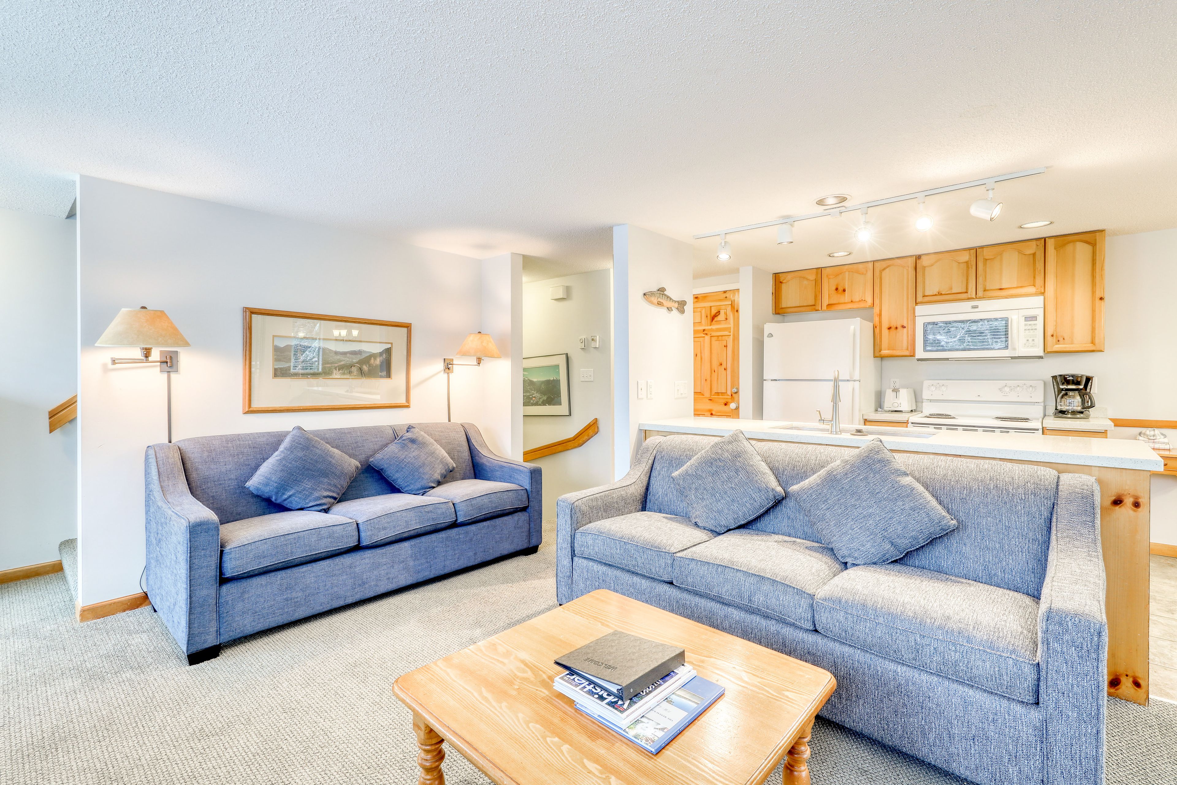 A modern living room with two sofas, a coffee table, and a kitchen area with wooden cabinets and appliances.