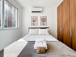 Quarto moderno com uma cama grande, roupa de cama branca, tapete cinza e janela com vista para edifícios.