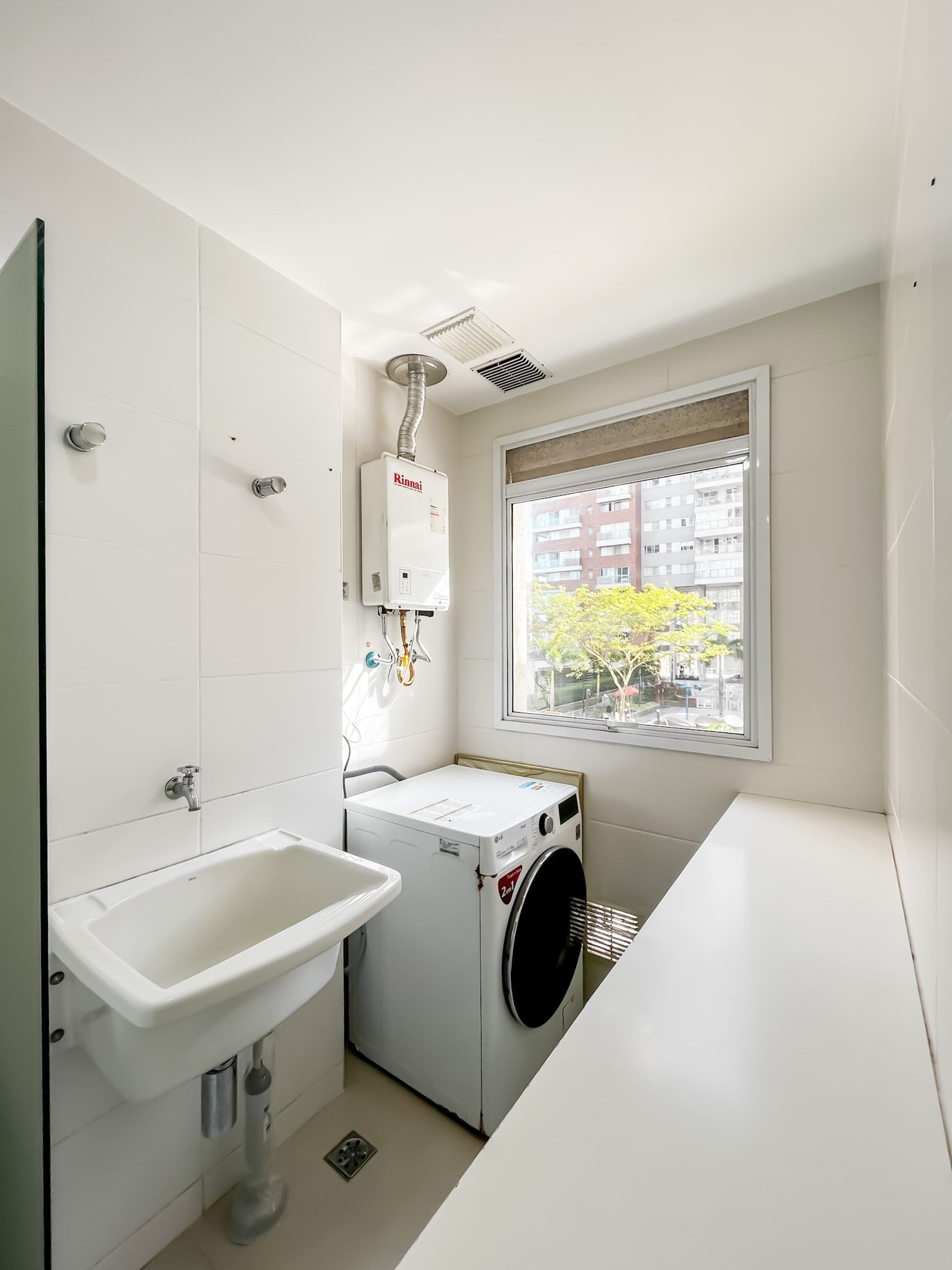 A compact laundry room with a washing machine, a sink, and a window with a view of buildings and trees.