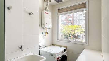 A compact laundry room with a washing machine, a sink, and a window with a view of buildings and trees.