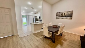 A dining area with a dark wooden table and white chairs, a kitchen in the background, and a painting on the wall. - Peaceful 3-bedroom house with AC in charming Palm Coast (Palm Coast)