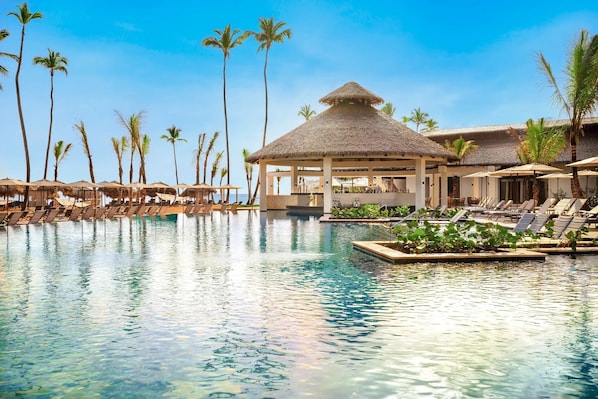 Ein Resort-Pool mit Liegestühlen, Palmen und einem Pavillon mit Reetdach.