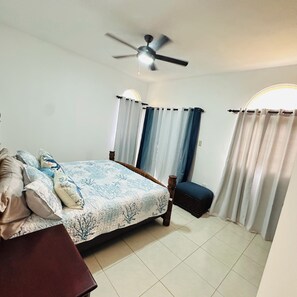Ein Schlafzimmer mit Bett, Nachttisch, Stuhl, Vorhängen, Deckenventilator und einem Fenster mit Vorhängen.