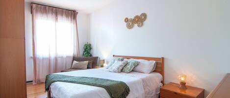 A bedroom with a wooden bed, a bedside table with a lamp, a window with curtains, and a wall decoration.