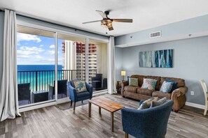 A spacious balcony with a view of the ocean and buildings, a ceiling fan, a brown leather sofa, and a wooden coffee table. - Beachfront 2BR+Bunks | Gulf View & Lagoon Pool (Panama City Beach)