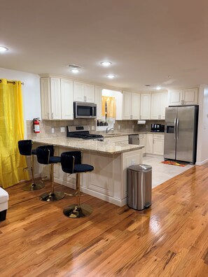 A modern kitchen with white cabinetry, a central island with bar stools, stainless steel appliances, and a yellow curtain.