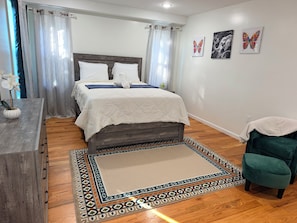 Un dormitorio con cama, mesita de noche, silla, alfombra y una obra de arte en la pared.