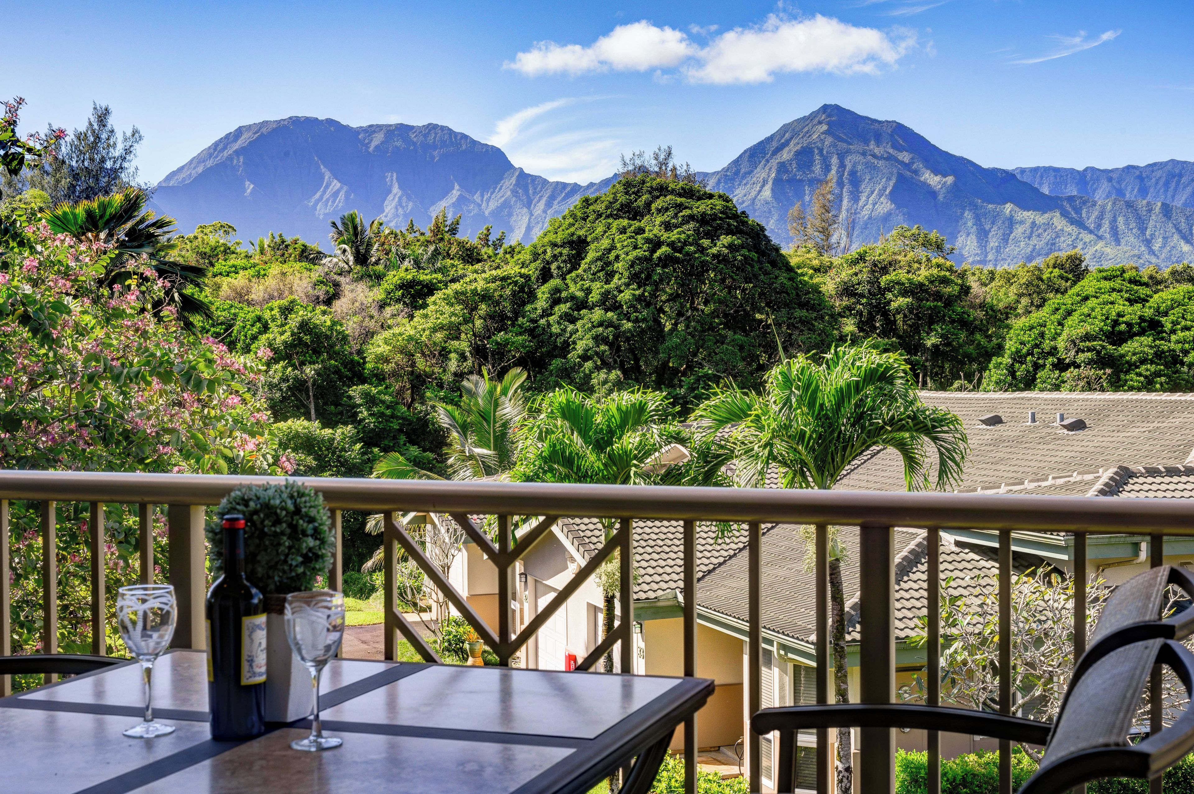 A terrace with a table, two wine glasses, and a bottle of wine. The terrace overlooks a mountainous landscape with lush greenery.