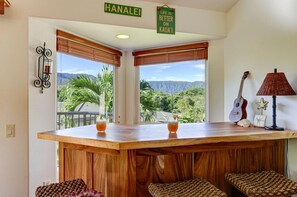 A bar area with a wooden counter, two glasses of a beverage, a guitar, and a sign that reads 'HA'NALEI'. - BEAUTIFUL HAWAIIANA 3/3 TOWNHOUSE WITH AC & MOUNTAIN VIEWS (Princeville)