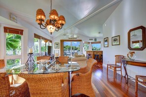 A spacious room with a glass dining table, wicker chairs, a chandelier, and large windows offering a view of greenery. - BEAUTIFUL HAWAIIANA 3/3 TOWNHOUSE WITH AC & MOUNTAIN VIEWS (Princeville)