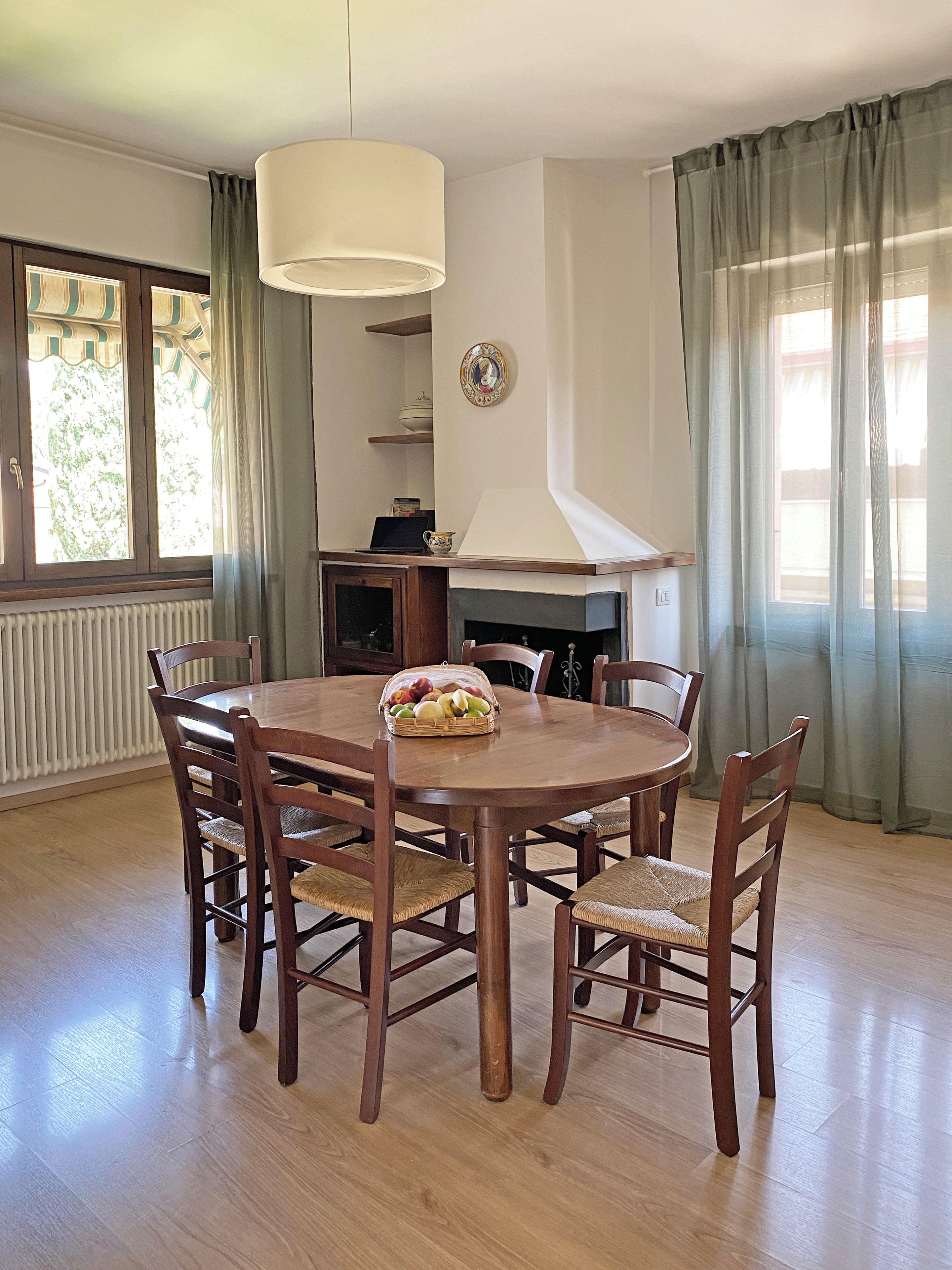 A dining room with a round wooden table, six chairs, a chandelier, a fireplace, and a window with curtains.
