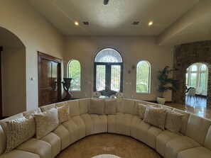 A spacious living room with a curved sectional sofa, large windows, and a stone wall. - Luxury Hill Country Estate - Sleeps 18 - Perfect for Groups (New Braunfels)