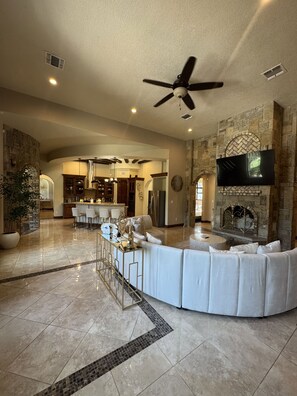 A spacious living area with a curved white sofa, a flat-screen TV mounted on a stone wall, and a stone fireplace. - Luxury Hill Country Estate - Sleeps 18 - Perfect for Groups (New Braunfels)